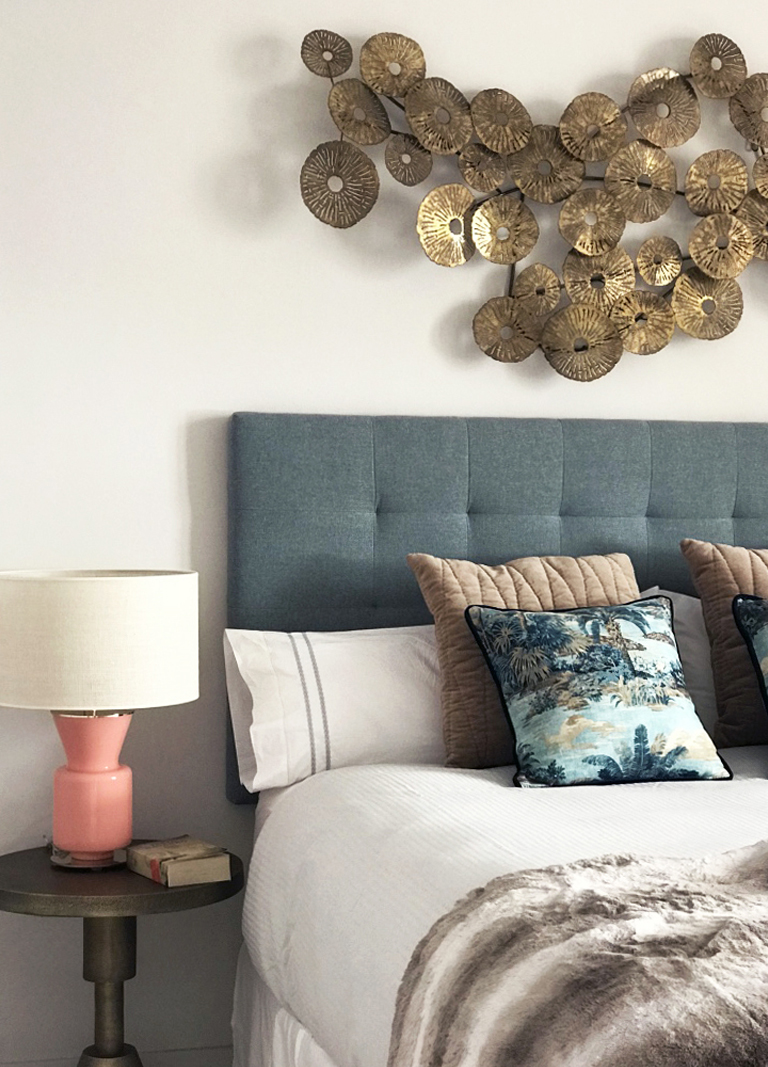 Habitación principal con cabecero en tela gris, aplique decorativo dorado en pared, mesilla de noche metalizada y lámpara sobre mesa rosada con pantalla blanca.