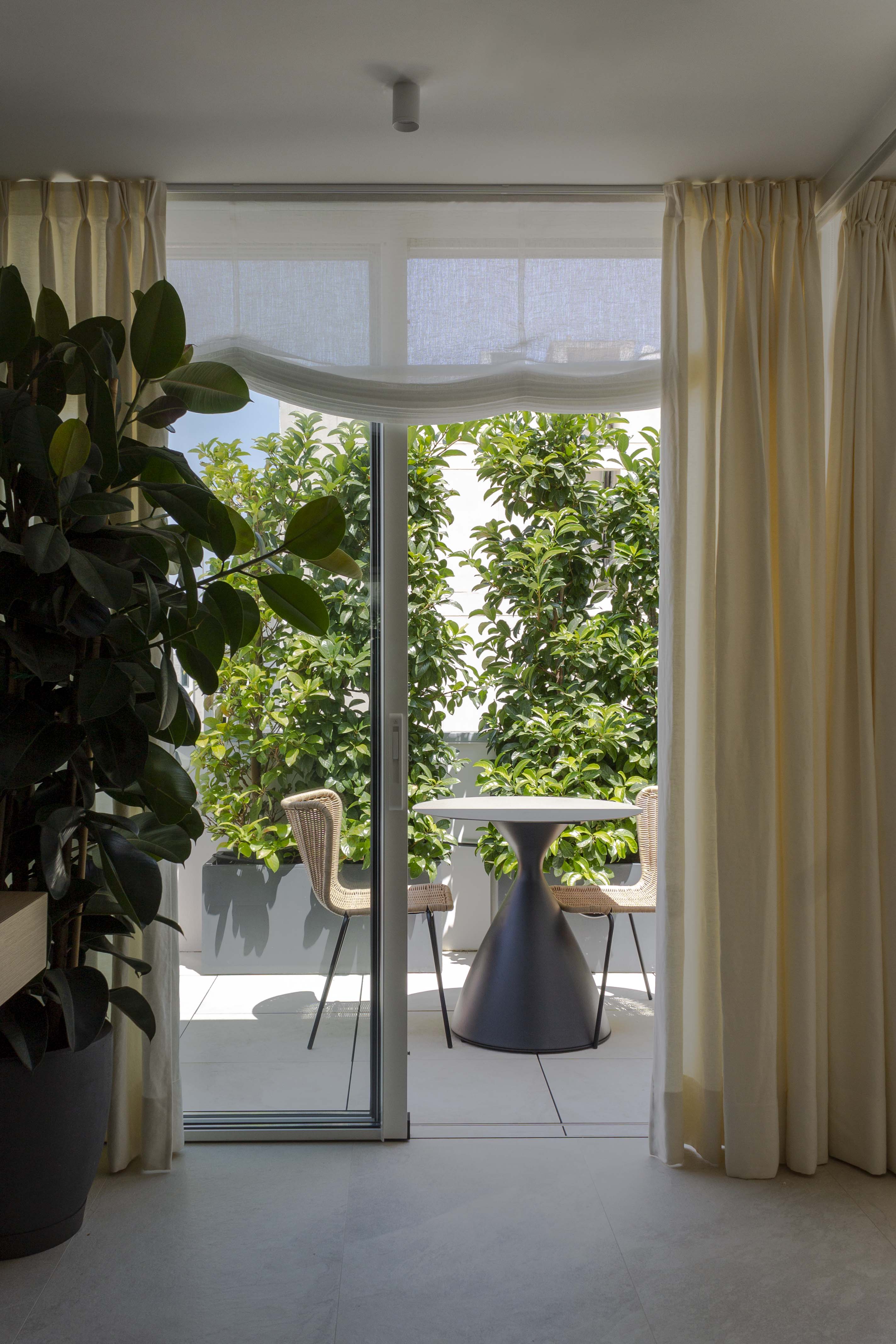 Vista de terraza desde despacho, decorada con mobiliario para exterior y vegetación alta para más privacidad. Cortinas de suelo a techo y estores