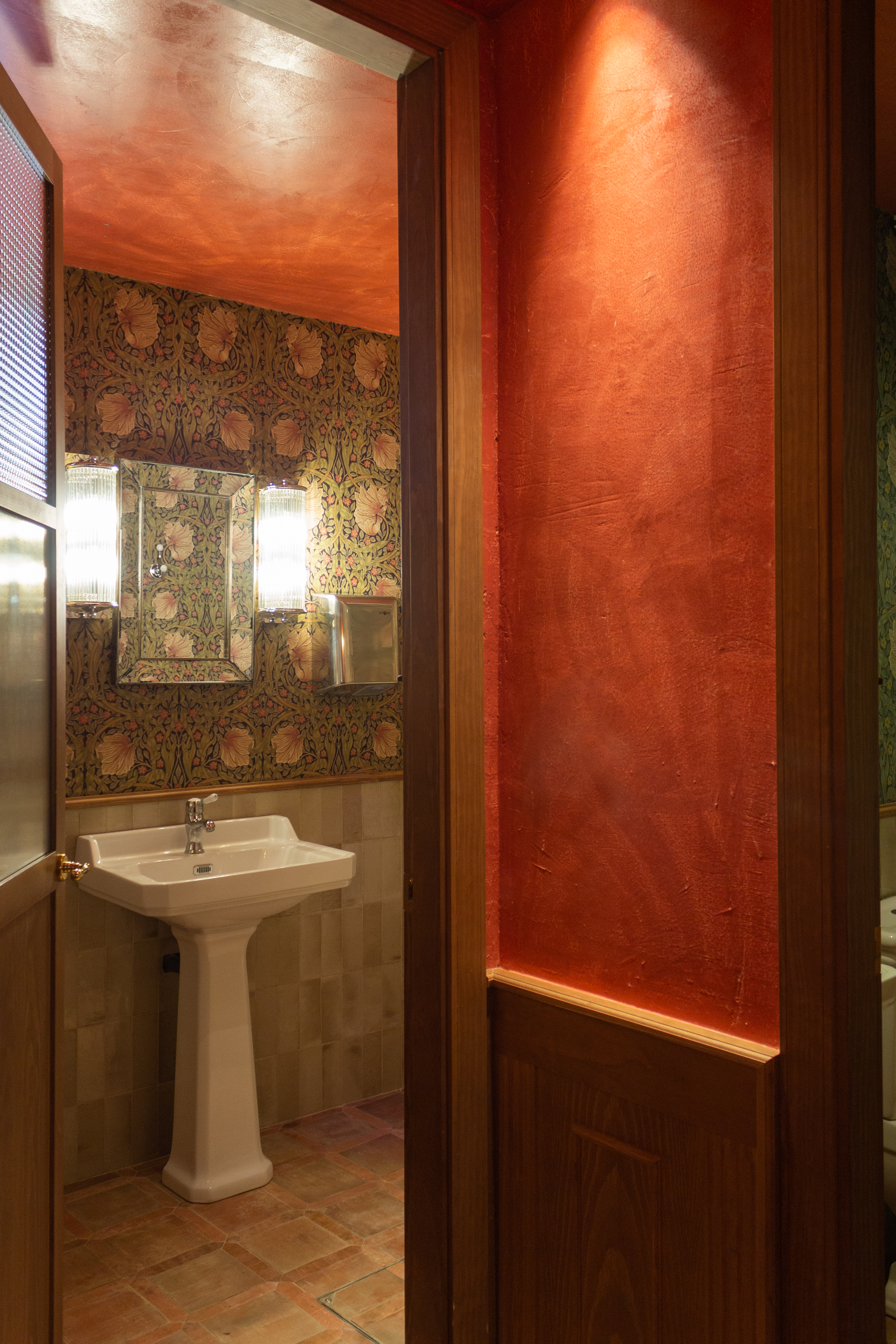 Vista al baño de señoras, diseñado con zócalo de alicatado a media altura y papel pintado llamativo, con un toque antiguo, clásico y elegante